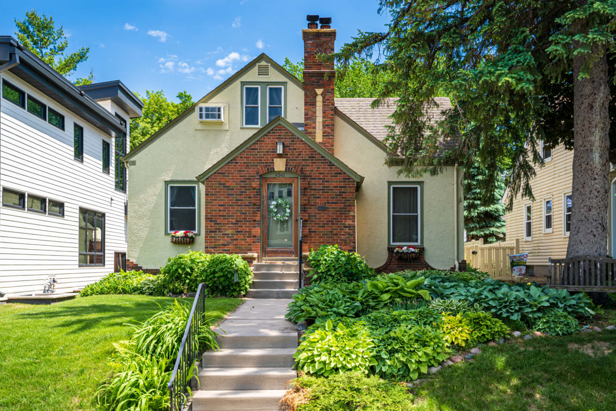 Charming brick and stucco home featuring a welcoming front entrance surrounded by mature landscaping in a quiet residential neighborhood.