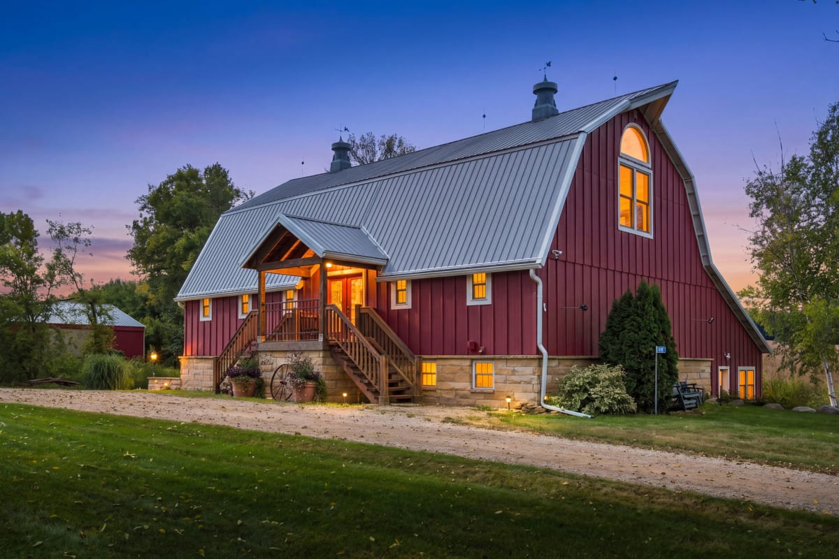 A beautifully restored barn-style property bathed in warm twilight, featuring charming red siding, rustic stone accents, and a welcoming covered entry—perfect for a peaceful countryside retreat.