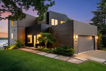 A contemporary lakeside home with black and wood panels, illuminated entrance, and landscaped path, under a twilight sky.