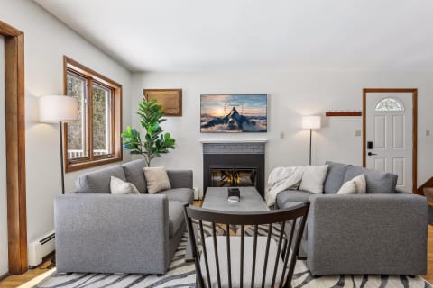 A modern living room featuring gray sofas, a fireplace, a mountain painting, and large windows.