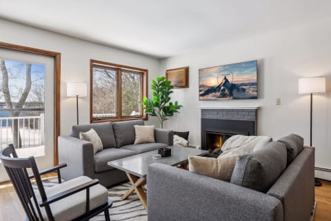 A bright living room featuring grey sofas, a metallic coffee table, a fireplace, and a view of trees and water outside the window.