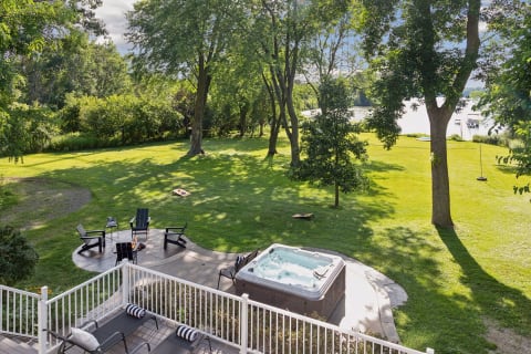 A spacious backyard with a hot tub, fire pit area, and open lawn bordered by trees, leading to a serene lake.