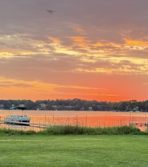 A sunset over a lake, featuring brilliant colors and a docked pontoon boat amidst lush green grass.