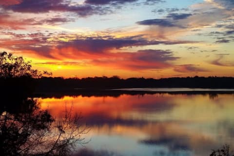 Sunset with vibrant hues of orange, pink, and purple reflected in a calm lake, framed by silhouetted trees.