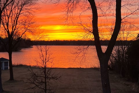 A vibrant sunset over a calm body of water, bordered by bare trees and a small structure.