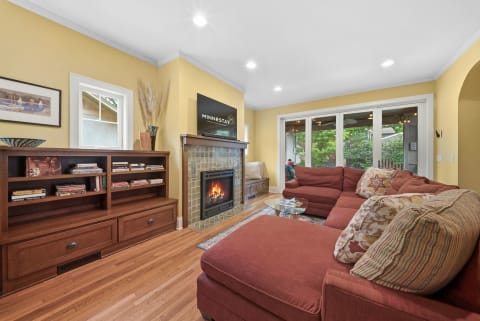 A cozy living room with a rust-colored sectional sofa, a fireplace, and large windows overlooking greenery.