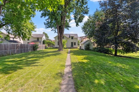 A green backyard featuring a stone pathway, tall trees, and two houses in a suburban setting.