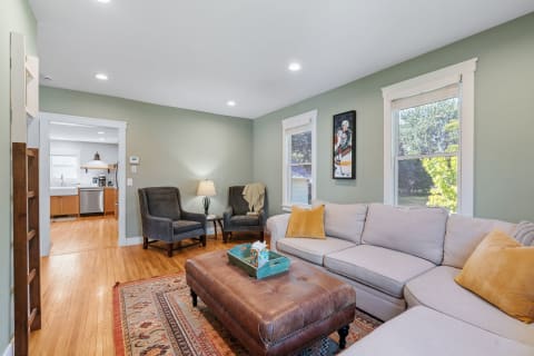 A living room featuring a beige sectional sofa, gray armchairs, and a leather ottoman, with an open view to a kitchen.