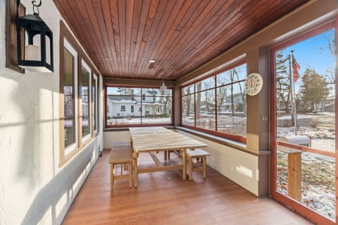 A bright sunroom with a wooden table and bench seating, surrounded by large windows and a warm wood ceiling.