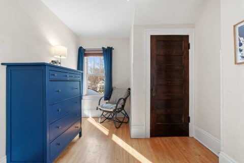 A corner of a room with a blue dresser, a chair, a window with blue curtains, and a wooden door.