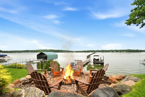 A fire pit surrounded by Adirondack chairs next to a tranquil lake, with boat docks in the background.
