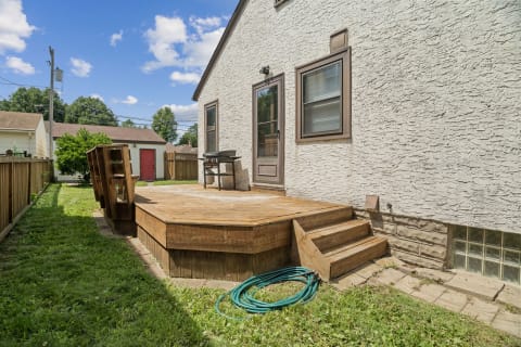 A wooden deck in a backyard featuring a grill, surrounded by green grass and a fence under a blue sky.