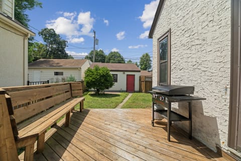 A wooden deck with a bench and grill overlooks a green yard with a shed and vibrant trees under a clear blue sky.