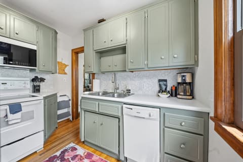 A modern kitchen with sage-green cabinetry, white countertops, and wooden floors, featuring a dual sink and a coffee maker.