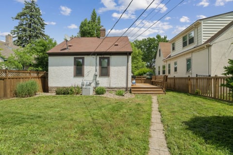A quaint white cottage in a backyard surrounded by green grass and trees beneath a blue sky.