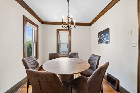 Cozy dining room with round table and brown chairs, brightened by a chandelier and natural light.