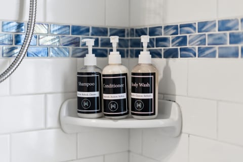 Three labeled bottles of shampoo, conditioner, and body wash on a white shelf in a shower, with blue tiles in the background.
