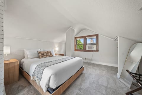 A cozy attic bedroom featuring a queen-sized bed, vaulted ceilings, and natural light from large windows.