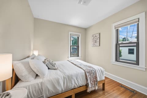 A bright bedroom featuring a stylish bed, warm wood floors, and natural light streaming through windows.