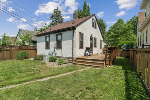 A cozy cottage backyard featuring a wooden deck, green lawn, and trees under a blue sky.