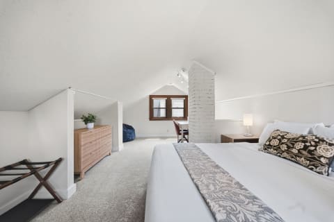 A serene attic bedroom with a bed, wooden dresser, bean bag, and cozy decor, illuminated by natural light.