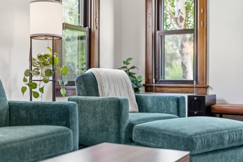 A cozy lounge area with teal velvet sofas, a warm lamp, and a houseplant near large windows.