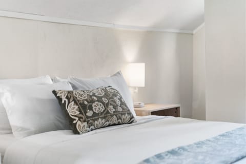 A tranquil bedroom featuring plush white linens, decorative pillows, a bedside lamp, and a wooden nightstand.