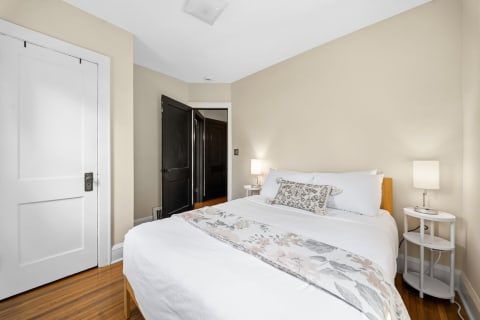 A cozy bedroom featuring a double bed with white linens, a floral throw, and a bedside lamp on a small table.