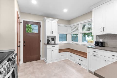 Modern kitchen with white cabinets, a wooden bench, and large windows.