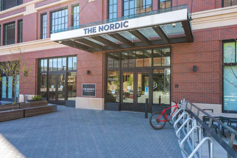 The entrance of The Nordic building with a brick facade, glass doors, bike storage, and planters.