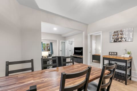Interior view of a dining area with a wooden table, black chairs, and a glimpse of a living room beyond.