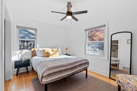 A cozy bedroom featuring a bed, nightstand, mirror, and colorful chair with autumn trees outside the window.