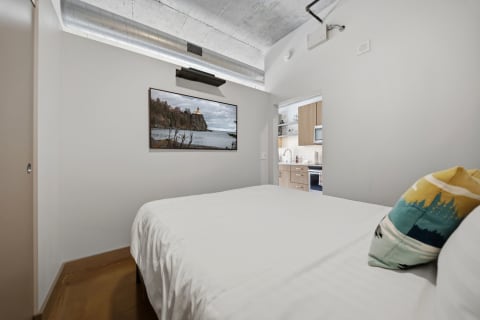 A modern bedroom with white bedding and a colorful decorative pillow, showcasing a scenic photograph on the wall.