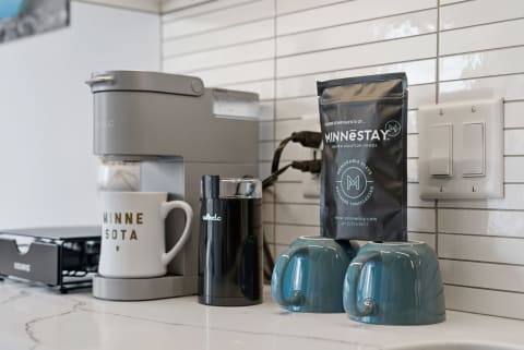 A modern coffee brewing setup with a white mug, black grinder, turquoise ceramic mugs, and coffee bean bag against a tiled wall.