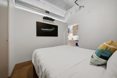 A cozy bedroom with a white bed, a wall-mounted TV, and access to a kitchen area in the background.