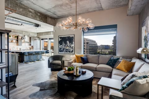 Interior of a contemporary living room with a kitchen and city views through large windows.