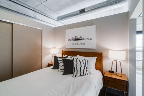 A cozy bedroom with a bed, geometric pillows, a nightstand with a lamp, and a Minneapolis skyline artwork on the wall.