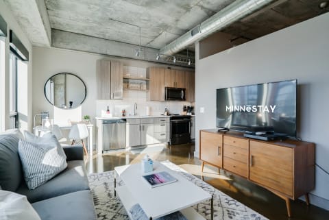 A stylish, modern apartment featuring a gray sofa, kitchen, and flat-screen TV.