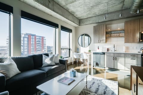 Modern apartment interior with a gray sofa, kitchen, and large windows.