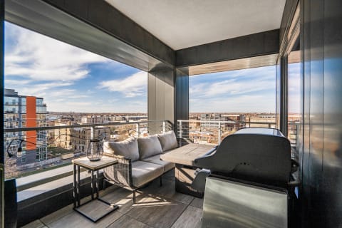 An urban balcony with a sofa, table, and grill overlooking the city skyline.