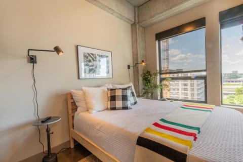Modern bedroom with a white bedspread, colorful throw, and decorative pillows, featuring large windows and a plant.