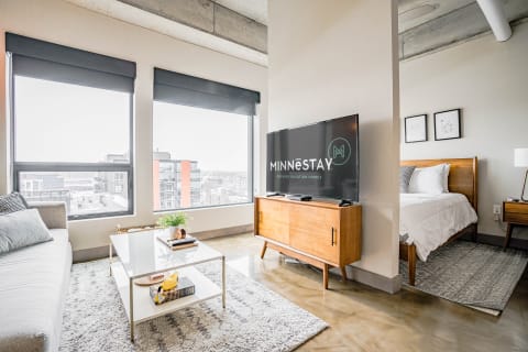 A stylish living area with large windows, a gray sofa, and a bedroom nook.