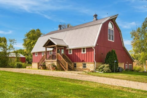 A charming red barn-style house in the countryside with a curved roof, stone foundation, and wooden stairs.