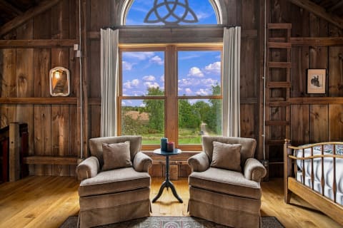 A rustic wooden room with plush chairs and a scenic view of the countryside through large windows.