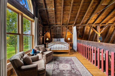 A rustic loft bedroom with wooden beams, a bed, and seating area, showcasing large windows with a view of greenery.