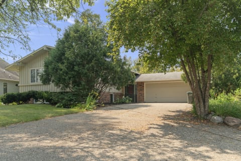A split-level home with green siding and a brick facade, featuring a gravel driveway and mature trees.