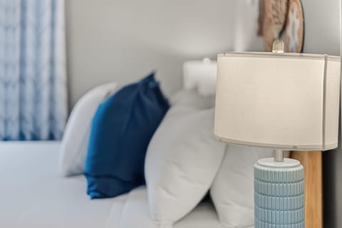 A cozy bedroom with a lamp, blue pillows, and white linens, reflecting a coastal theme.