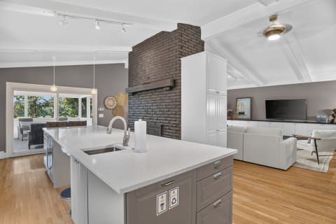 A modern kitchen with a white island, gray cabinetry, and a black brick fireplace, connecting to a living area with a large TV.