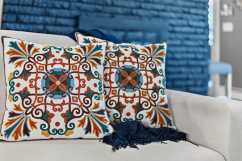Two vibrant embroidered pillows on a beige couch with a blue textured wall in the background.