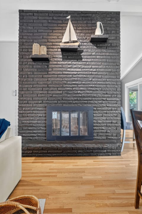 A cozy living room featuring a gray brick fireplace with nautical decor on shelves, surrounded by warm wooden flooring and a white sofa with a blue throw.